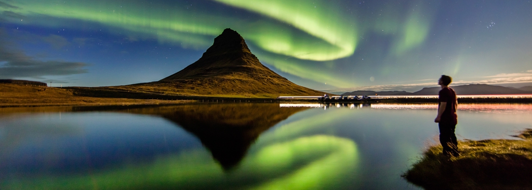 Man standing on edge of shore looking at body of water with northern lights reflecting in lake