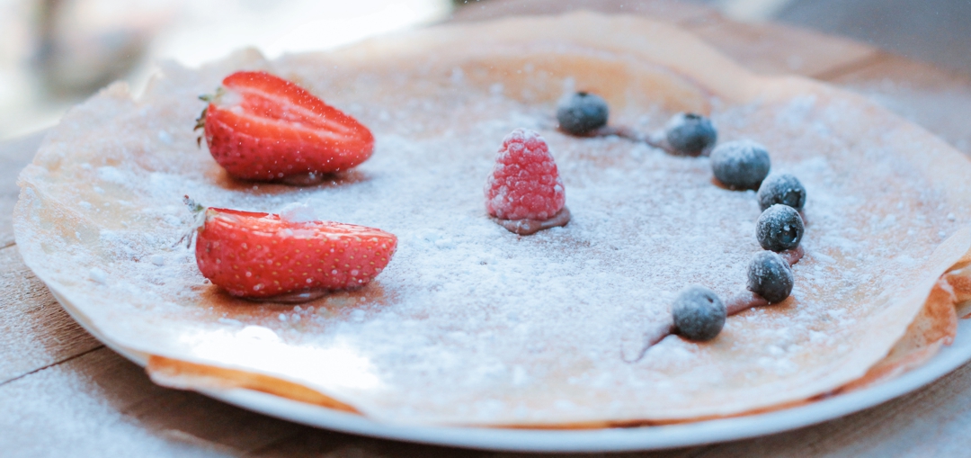 crepe on dinner plate with smiley face made from strawberry slices and blueberries and powdered sugar