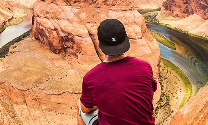 young man with back to camera sitting at edge of Grand Canyon with Colorado River below