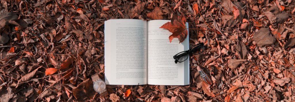 open book outdoors lying in fallen autumn leaves with a pair of glasses and wrist watch