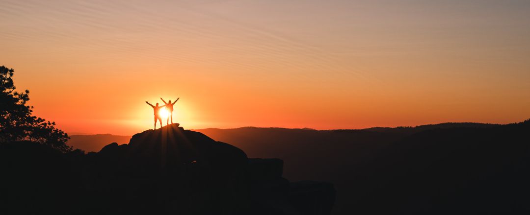 two people standing with arms raised at the top of a knoll silhouetted against the sun