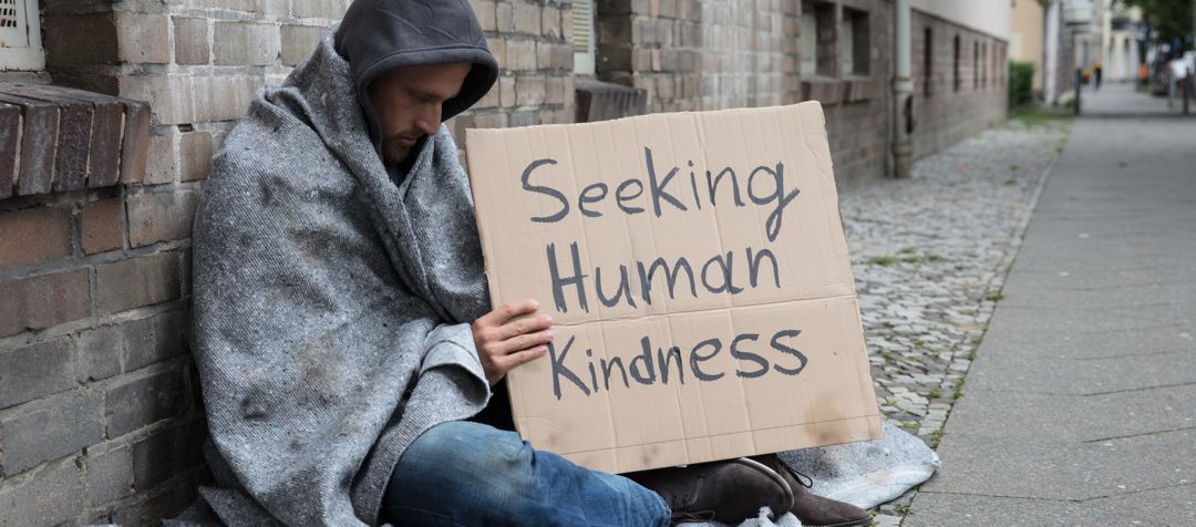 Homeless man sitting on ground leaning against brick wall holding a sign that says "seeking human kindness"