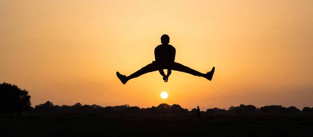 male figure jumping with legs splayed out to side, pointing to sunrise