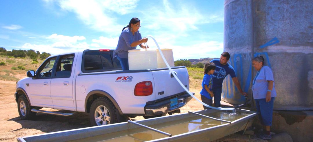 Two women, 1 man, and 1 child filing large water vessel from water tank