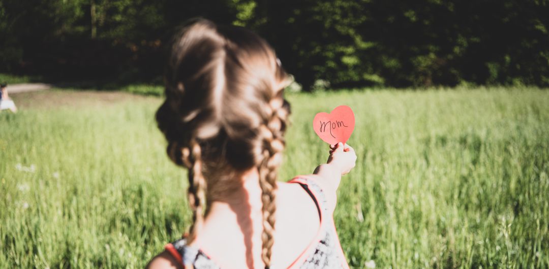 little girl with braided pigtails standing in field with outstretched arm holding red paper heart with the word "mom" on it