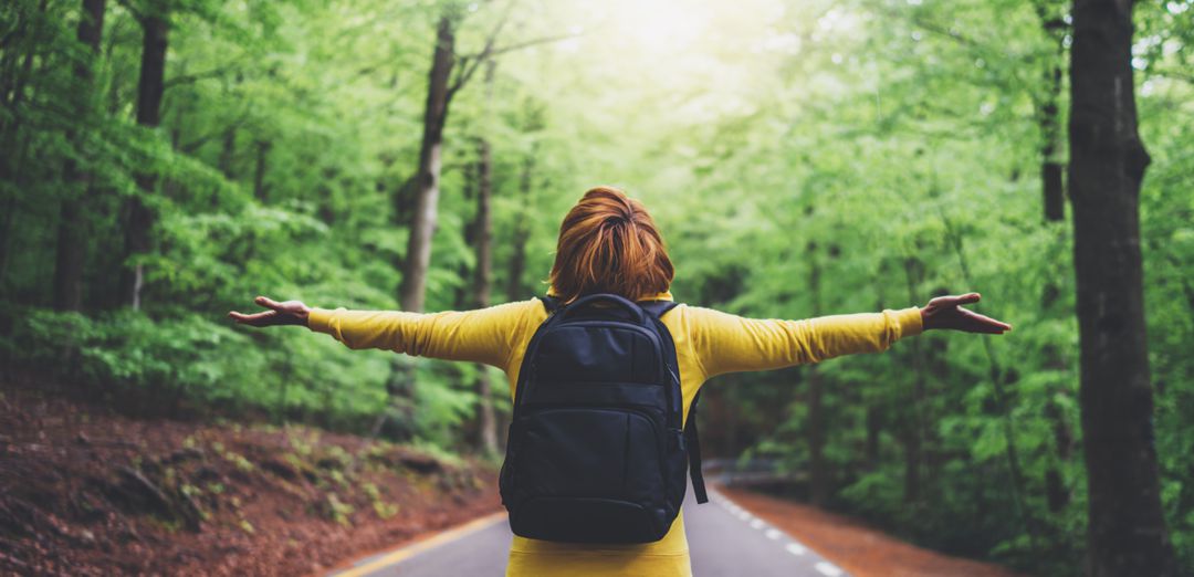 Woman standing in the middle of the road in forest wearing a backpack with arms spread wide breathing fresh air