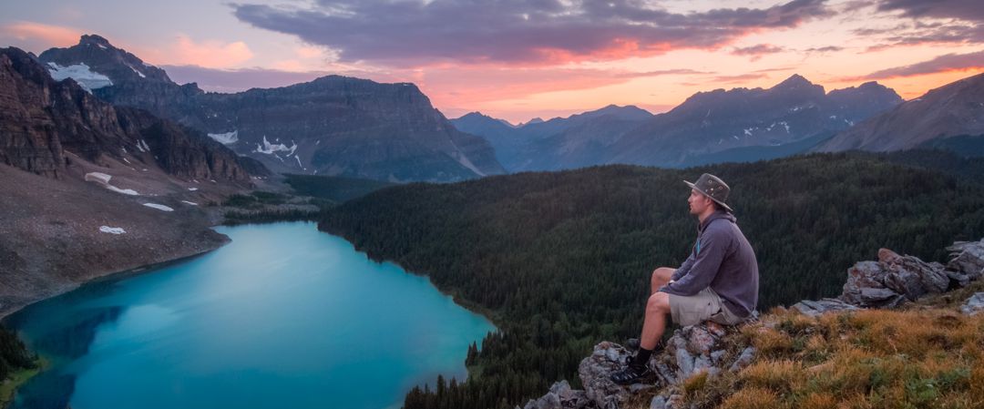 man sitting at the top of a mountain looking looking across vast scenery consisting of lake, forest, and mountains