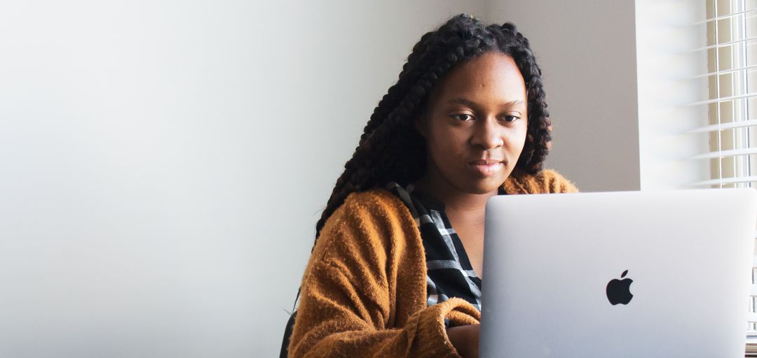 Woman of color sitting next to window in front of an Apple computer