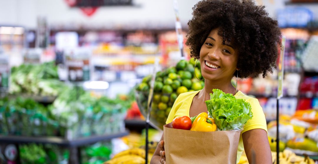 Healthy positive happy woman of color holding a paper shopping bag full of fruit and vegetables.