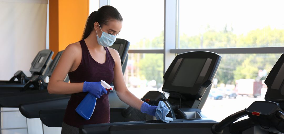 Woman cleaning treadmill with disinfectant spray and cloth in gym